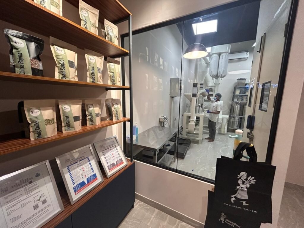 The glass-walled milling room at Slomilled Foods, M3M Urbana, with a miller at the stone chakki and packed pulses on the shelves outside.
