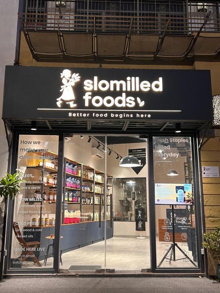 The Slomilled Foods storefront at night, with an illuminated sign reading “Better food begins here” and window graphics explaining how staples are made.