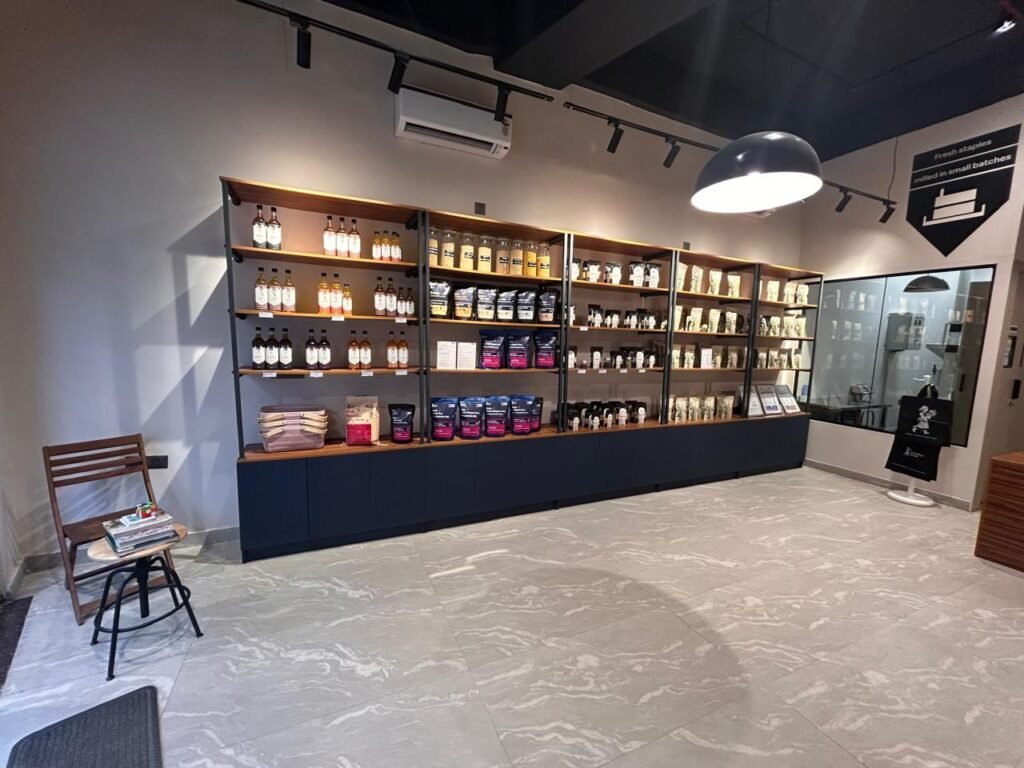 Wood and steel shelving at Slomilled holding bottled oils, jars of grains and packed flours, with the milling room window on the right.